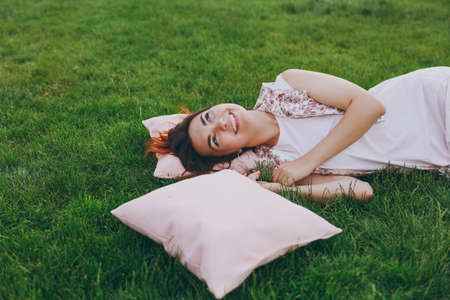 Cose up portrait of smiling happy joyful beautiful woman in light dress lying on pink pillow on green grass lawn in city park, rest, relax, have fun and looking up. Young female with ginger hairの写真素材