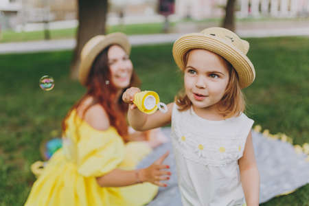 Woman in yellow clothes play on grass in park rest, amuse with little cute child baby girl blowing soap bubbles. Mother, little kid daughter. Mother's Day, love family, parenthood, childhood conceptの写真素材