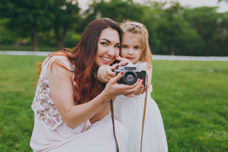 Pretty woman in light dress and little cute child baby girl take picture on retro vintage photo camera in park. Mother, little kid daughter. Mother's Day, love family, parenthood, childhood conceptの写真素材
