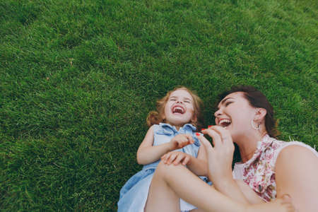 Overjoyed woman in light dress and little cute child baby girl lie on green grass in park rest play and have fun. Mother, little kid daughter. Mother's Day, love family, parenthood, childhood conceptの写真素材