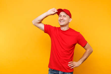 Delivery man in red uniform isolated on yellow orange background. Professional smiling male employee in t-shirt working as courier or dealer put hand on cap head gesture. Service concept.の写真素材
