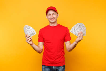 Delivery man in red uniform isolated on yellow orange background. Professional male employee in cap, t-shirt courier or dealer holding bundle lots of dollars, cash money.の写真素材