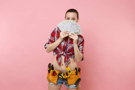 Young handyman woman in plaid shirt, denim shorts, kit tools belt full of variety instruments hold...