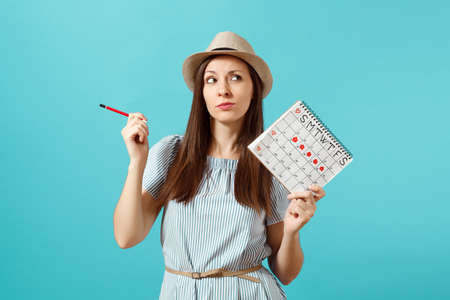 Portrait pensive woman in blue dress, hat holding red pencil, female periods calendar for checking menstruation days isolated on blue background. Medical healthcare, gynecological concept.の写真素材