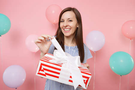 Portrait of smiling young woman in blue dress untying bow on red box with gift present on pastel...