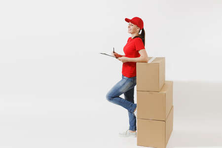 Full length of delivery woman in red cap, t-shirt isolated on white background. Female courier holding clipboard with papers document, blank empty sheet on empty cardboard boxes. Receiving packageの写真素材