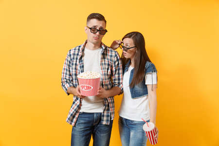 Young exhausted couple woman and man in 3d glasses watching movie film on date holding bucket of popcorn and plastic cup of soda or cola isolated on yellow background. Emotions in cinema conceptの写真素材