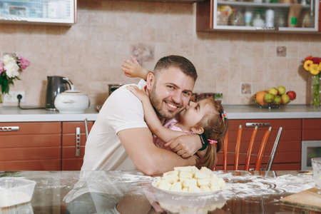 Little kid girl help man to cook lazy dumplings kissing daddy in kitchen at table. Happy family dad, child daughter cooking food in weekend morning at home. Father's day holiday. Parenthood childhoodの写真素材