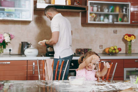 Little kid girl plays with flour while daddy cooking in kitchen at table. Happy family dad, child...