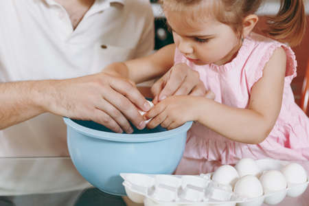 Close up Little kid girl help man to cook Christmas ginger cookies, breaks egg into bowl at table....