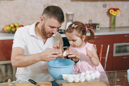 Little kid girl helps man to cook Christmas ginger cookies, breaks egg into bowl at table. Happy...
