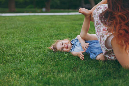 Woman in light dress and smiling little cute child baby girl lie on green grass in park rest play and have fun. Mother, little kid daughter. Mother's Day, love family, parenthood, childhood conceptの写真素材