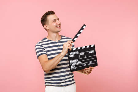 Portrait of young man wearing striped t-shirt holding classic black film making clapperboard isolated on trending pastel pink background. People sincere emotions lifestyle concept. Advertising areaの写真素材