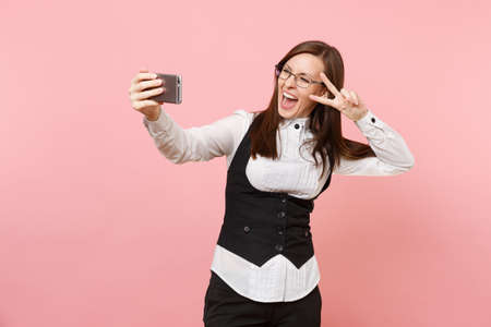 Young overjoyed business woman in glasses doing taking selfie shot on mobile phone showing victory sign isolated on pink background. Lady boss. Achievement career wealth. Copy space for advertisementの写真素材