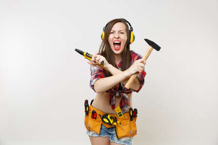 Angry emotional woman in tartan shirt, denim shorts, noise insulated headphones, kit tools belt full of instruments isolated on white background. Female in male work. Renovation occupation conceptの写真素材