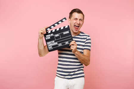 Portrait of young man wearing striped t-shirt holding classic black film making clapperboard isolated on trending pastel pink background. People sincere emotions lifestyle concept. Advertising areaの写真素材