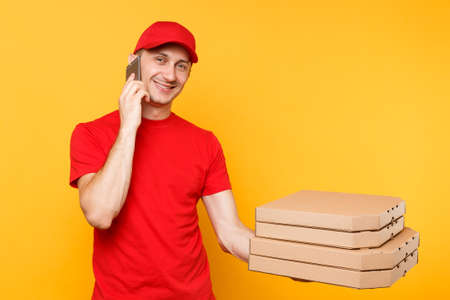 Delivery man in red cap t-shirt giving food order italian pizza in blank empty flatbox boxes on yellow background. Male employee pizzaman courier talking on mobile cell phone mockup. Service conceptの写真素材