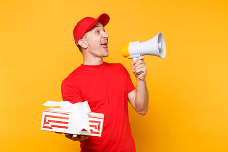 Delivery service man in red uniform isolated on yellow background. Male employee courier in cap, t-shirt screaming in megaphone hot news hold gift box. Fun guy announces discounts sale. Copy spaceの写真素材
