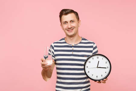 Portrait of happy caucasian man in striped t-shirt holding round clock, clear fresh pure water in...