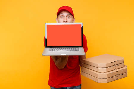 Delivery man in red cap t-shirt giving food order pizza in flatbox boxes on yellow background. Male employee pizzaman courier hold laptop pc computer with blank empty screen mockup. Service conceptの写真素材