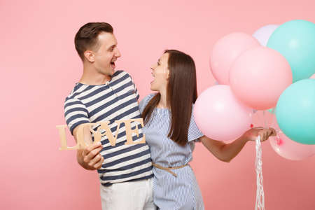 Portrait couple hold wooden word letters love. Woman and man in blue clothes celebrating birthday holiday party on pastel pink background with colorful air balloons. People sincere emotions conceptの写真素材