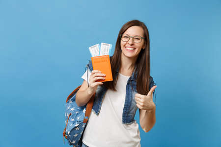 Young happy woman student in glasses with backpack holding passport, boarding pass tickets and showing thumb up isolated on blue background. Education in university college abroad. Air travel flightの写真素材