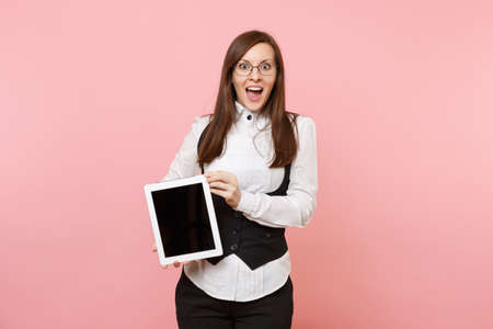 Young shocked amazed business woman in suit and glasses holding tablet pc computer with blank empty screen isolated on pastel pink background. Lady boss. Achievement career wealth concept. Copy spaceの写真素材