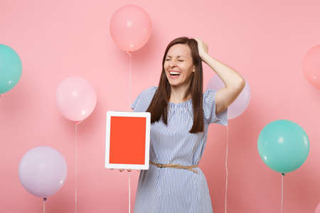 Portrait of laughing young woman in blue dress holding tablet pc computer with blank empty screen clinging to head on pastel pink background with colorful air balloons. Birthday holiday party conceptの写真素材