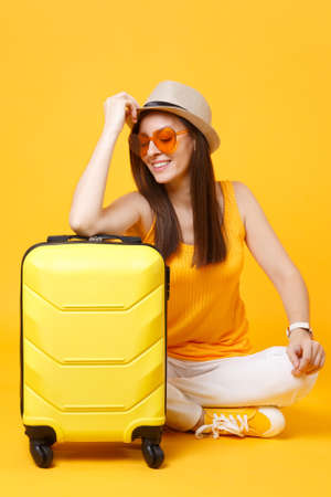 Traveler tourist woman in summer casual clothes, hat sit with suitcase isolated on yellow orange background. Female passenger traveling abroad to travel on weekends getaway. Air journey conceptの写真素材