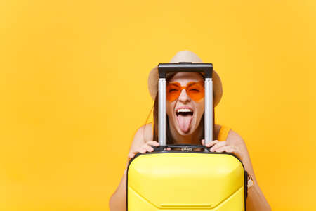 Fun traveler tourist woman in summer casual clothes, hat with suitcase showing tongue isolated on yellow background. Passenger traveling abroad to travel on weekends getaway. Air journey conceptの写真素材