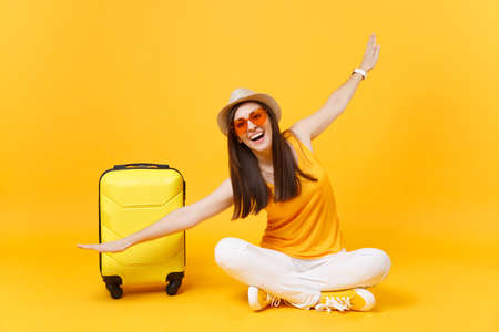 Tourist woman in summer clothes, hat sit at suitcase crossed legs spreading hands as in flight isolated on yellow orange background. Passenger traveling abroad on weekend getaway. Air journey conceptの写真素材