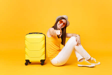 Traveler tourist woman in summer casual clothes, hat sit with suitcase isolated on yellow orange background. Female passenger traveling abroad to travel on weekends getaway. Air journey conceptの写真素材