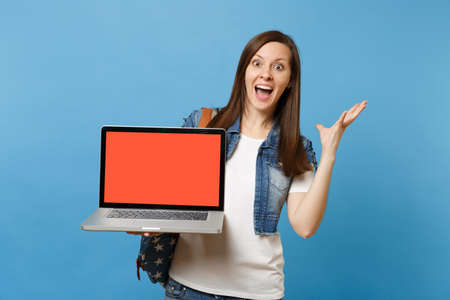 Young excited woman student with opened mouth spreading hands holding laptop pc computer with blank black empty screen isolated on blue background. Education in college. Copy space for advertisementの写真素材