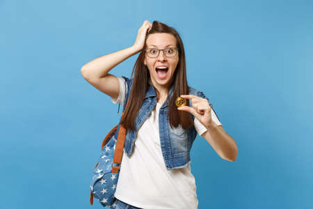 Young amazed shocked woman student with opened mouth clinging to head hold bitcoin metal coin of golden color isolated on blue background. Future currency. Education in high school university collegeの写真素材