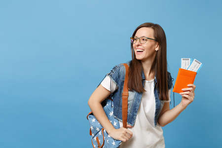 Young laughing woman student in glasses with backpack looking aside hold passport, boarding pass tickets isolated on blue background. Education in university college abroad. Air travel flight conceptの写真素材