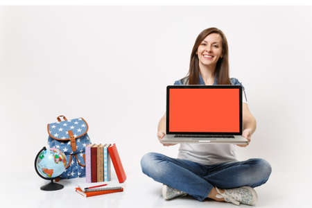 Young joyful woman student holding laptop pc computer with blank black empty screen sitting near globe, backpack school books isolated on white background. Education in high school university collegeの写真素材