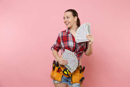 Young handyman woman in plaid shirt, denim shorts, kit tools belt full of variety instruments hold lots of cash dollar money isolated on pink background. Female doing male work. Renovation conceptの写真素材