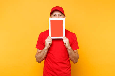 Delivery man isolated on yellow orange background. Professional male employee courier in red cap, t-shirt holding tablet computer with blank empty screen. Service concept. Copy space. Mockup templateの写真素材