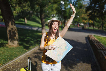 Excited traveler tourist woman with suitcase city map waving hand for greeting, meet friend, catch taxi in city outdoor. Girl traveling abroad to travel on weekends getaway. Tourism journey lifestyleの写真素材