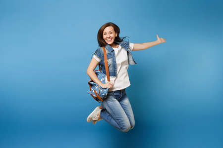 Full length portrait of young happy joyful woman student in denim clothes with backpack jumping and spreading hands isolated on blue background. Education in college. Copy space for advertisementの写真素材