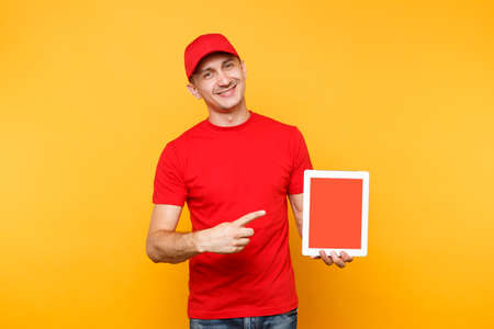 Delivery man isolated on yellow orange background. Professional male employee courier in red cap, t-shirt holding tablet computer with blank empty screen. Service concept. Copy space. Mockup templateの写真素材