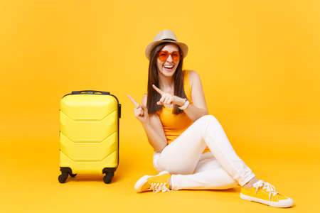 Traveler tourist woman in summer casual clothes, hat sit with suitcase isolated on yellow orange background. Female passenger traveling abroad to travel on weekends getaway. Air journey conceptの写真素材