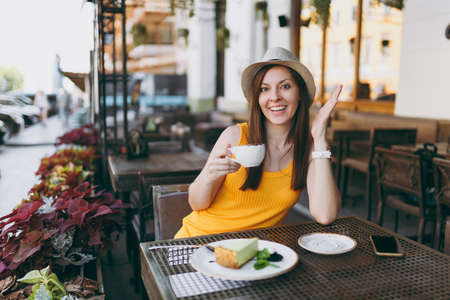 Woman in outdoors street coffee shop cafe sitting at table in yellow clothes hat with cup of cappuccino, cake, relaxing in restaurant during free time. Mobile Office in summer. Lifestyle rest conceptの写真素材