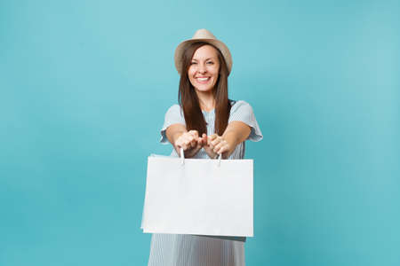 Portrait excited smiling beautiful caucasian woman in summer dress, straw hat holding packages bags with purchases after shopping isolated on blue pastel background. Copy space for advertisementの写真素材