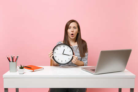 Young shocked puzzled woman holding round alarm clock while sit work at office with pc laptop isolated on pastel pink background. Achievement business career concept. Copy space. Time is running outの写真素材