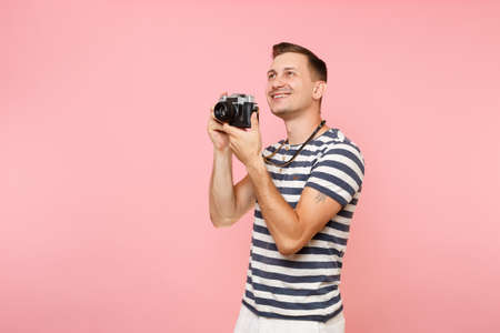 Portrait of smiling young photographer man wearing striped t-shirt take pictures on retro vintage photo camera isolated on trending pastel pink background. People sincere emotions lifestyle conceptの写真素材