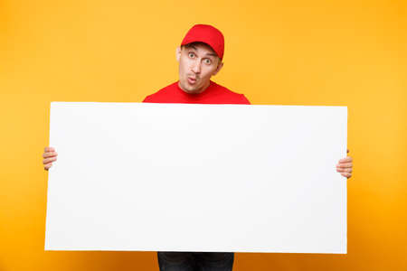 Delivery man in red uniform isolated on yellow orange background. Male employee courier in cap, t-shirt holding big white empty blank billboard with copy space, place for text image. Service conceptの写真素材