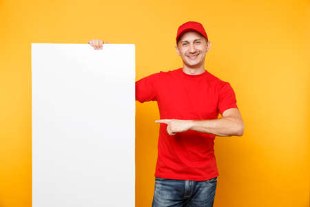 Delivery man in red uniform isolated on yellow orange background. Male employee courier in cap, t-shirt holding big white empty blank billboard with copy space, place for text image. Service conceptの写真素材