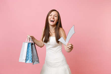 Happy bride woman in wedding dress holding check mark, multi colored packages bags with purchases after shopping isolated on pink background. Wedding to do list. Organization of celebration conceptの写真素材