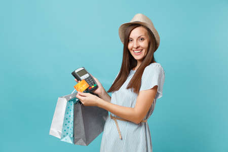 Portrait woman in dress, hat holding packages bags with purchases after shopping, wireless modern bank payment terminal to process and acquire credit card payments isolated on blue pastel backgroundの写真素材